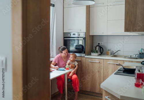 Mother feeds her baby in modern kitchen, surrounded by cozy, light-filled environment. Fresh fruits and potted plants decorate windowsill, adding warm, homely touch. Motherness warm concept photo.