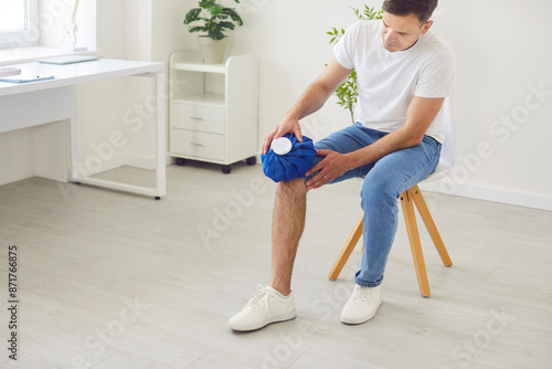 Portrait of a young man applying cold compress to leg sitting on chair at home. Male person holding ice pack bag on the knee to reduce pain. Inflammation, injury and health problems concept.