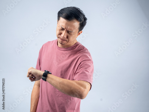 An Asian man in a pink shirt looks at his watch with a concerned and impatient expression, suggesting he is worried about time. The plain light blue background emphasizes his focused