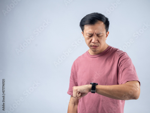 An Asian man in a pink shirt looks at his watch with a concerned and impatient expression, suggesting he is worried about time. The plain light blue background emphasizes his focused