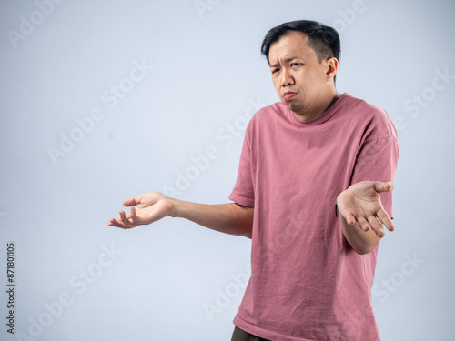 An Asian man in a pink shirt shrugs with a confused and uncertain expression, his arms outstretched and palms up. The plain light blue background emphasizes his ambiguous