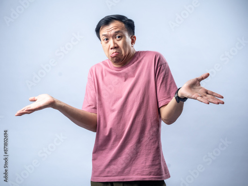 An Asian man in a pink shirt shrugs with a confused and uncertain expression, his arms outstretched and palms up. The plain light blue background emphasizes his ambiguous