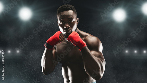 A boxer in red hand wraps stands ready in a dark stadium, illuminated by bright lights.