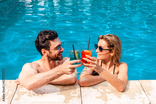 Smiling young woman and a cute dark-haired guy clinking glasses of cocktails in an outdoor swimming pool. Leisure and summer vacation concept