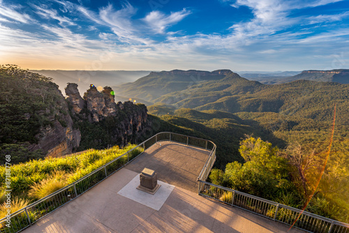 A panoramic view of the Three Sisters rock formation in the Blue Mountains, Australia. The photo was taken from the scenic Echo Point Lookout with a railing overlooking the vast landscape.