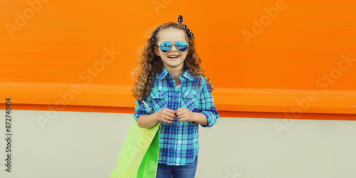 Stylish happy joyful kid girl standing with shopping bag, cheerful little girl child on city street