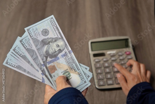 Close-up photo of a person's hands using a calculator and holding a wad of cash on a wooden table. Financial concept.