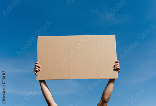 hands holding up a blank cardboard sign on clear blue sky background