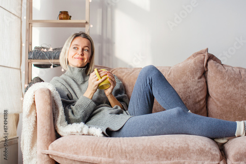 Happy dreamy middle aged woman sitting on sofa in living room with cup of tea or coffee