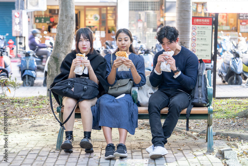 Three people - a Taiwanese woman, a Taiwanese man, and an Asian woman - sitting together on a bench in Da’an District, Taipei City, chatting and enjoying a large waffle.