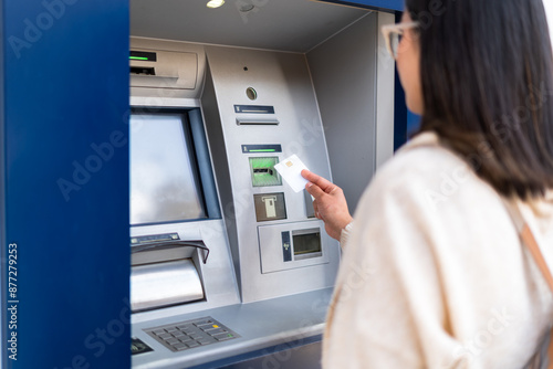 A woman uses an ATM machine, inserting her card for a transaction. The focus is on the card and machine. Finance and technology concept.