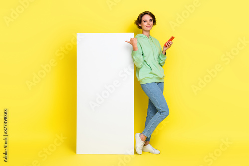 Full length photo of cute cheerful lady wear green hoodie typing modern gadget pointing thumb empty space isolated yellow color background