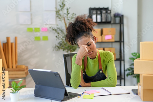 Young business owner is struggling with a headache while juggling her online business, surrounded by parcels and documents