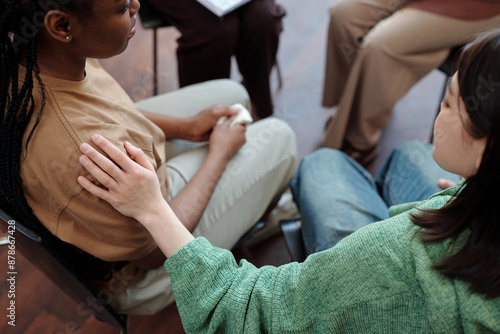 Young brunette woman in casual attire keeping hand on shoulder of upset African American girl while comforting her during session