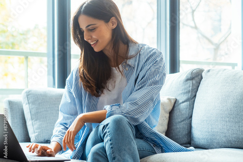 Relaxed woman using laptop in luxury home living room with big window, enjoying working, internet shopping, checking social network, reading news or communicating online with computer sitting on sofa