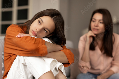 Depressed sad teenage girl sitting and hugging her knees, looking away while mother sitting next daughter. Communication problems between parent and kids