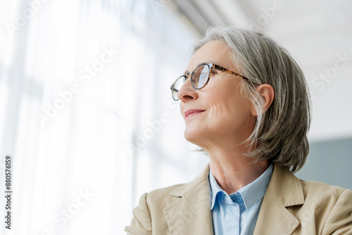 Senior businesswoman with eyeglasses smiling and looking up through office window, copy space