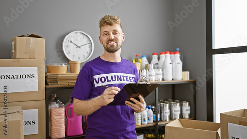 Smiling caucasian man with beard in volunteer shirt holding clipboard in a donation center warehouse.