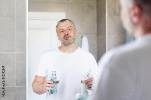 Middle aged man rinsing mouth with blue mouthwash, standing in bathroom in front of mirror. Teeth and oral care