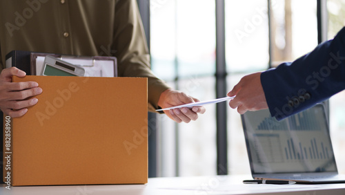 Two individuals exchanging documents across a desk. One person is holding a box filled with office supplies, indicating a possible job transition or project handover