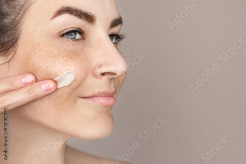 Caucasian woman's face with freckles or age spots, she smears cream on her cheek with her fingers, nice cream swatch, copy space for text