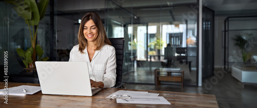 Middle age European business woman CEO using laptop application for work at table workspace in office. Smiling Latin Hispanic mature adult professional businesswoman using pc digital computer. Banner