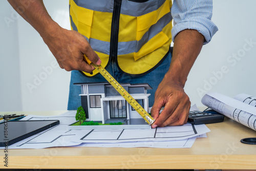 Asian architect works at a desk with house blueprints, checking plans on a laptop and clipboard, wearing a hardhat and safety vest, emphasizing precision and safety in office settings.