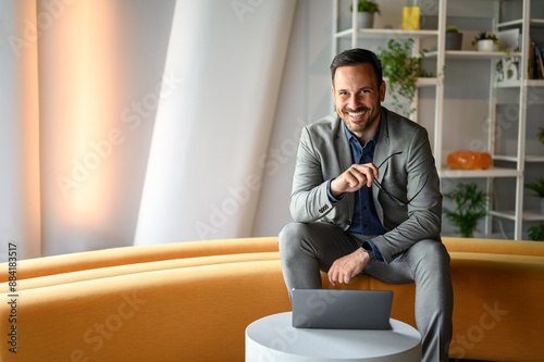 Portrait of handsome business leader holding glasses and smiling while working over laptop in office