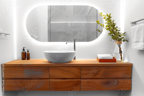 A minimalist mid-century modern bathroom featuring white walls, a floating wooden vanity with a vessel sink, and a large round mirror with LED backlighting.