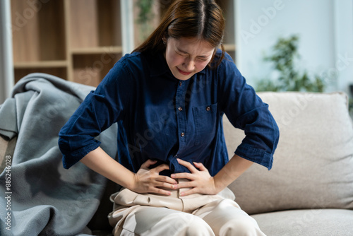 A young Asian woman sits on a sofa in her living room, feeling sick with a stomach ache. She experiences nausea, bloating, and cramping, seeking comfort and relief at home.