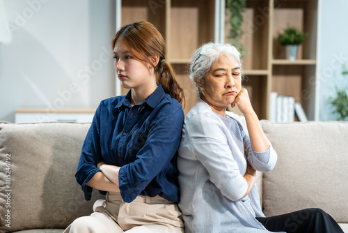 A mature mom and a young Asian woman, mother and daughter, sit together on a sofa in their living room, dealing with an argument and conflict, trying to resolve their differences at home.