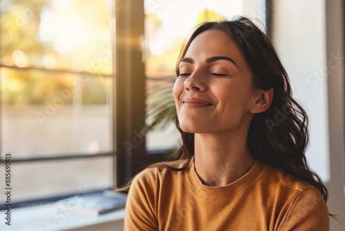 A woman with long dark hair, dressed in a mustard yellow sweater, sits by a window with her eyes closed, breathing deeply and basking in the warm sunlight