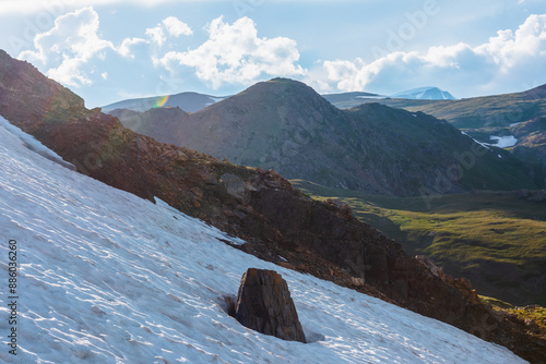 Slippery snow in bright sun under clouds in blue sky. Sunlit alpine landscape with diagonal snowfield with rock in center against big rocky hill, large mountain range and high snowy top in sunny day.