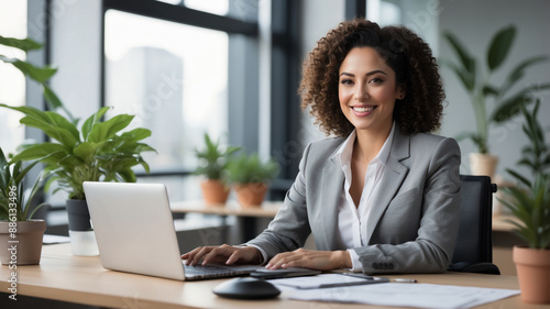 A businesswoman in a grey suit sits at her desk in a modern office, working on her laptop with a confident smile