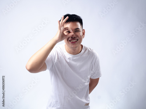 A young Asian man, wearing a white shirt and black watch, makes a playful expression while forming an "OK" sign around his eye with his hand. The plain white background highlights