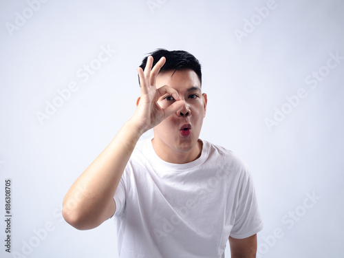 A young Asian man, wearing a white shirt and black watch, makes a playful expression while forming an "OK" sign around his eye with his hand. The plain white background highlights