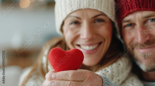 A close-up of a couple in winter hats and sweaters, smiling warmly while holding a small, red heart, radiating love, warmth, and happiness during a cozy indoor moment.