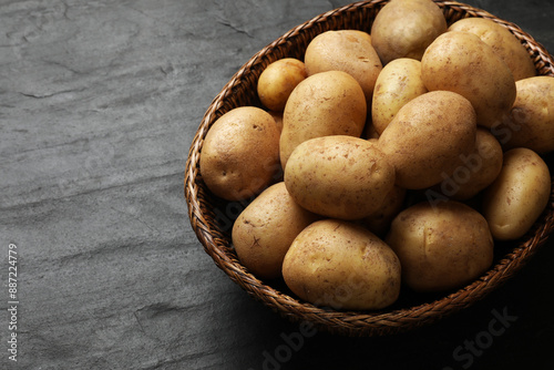 Many fresh potatoes in wicker basket on black textured table, closeup. Space for text