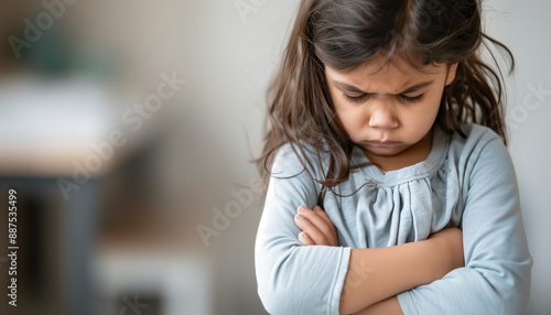 A young girl with long hair and a serious expression crosses her arms, conveying the emotion of sadness and frustration indoors.