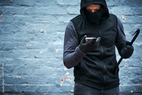 Thief, phone and crime outdoor for danger, illegal and burglar with hoodie, mask and steel bar as weapon. Criminal, smartphone and warning by wall for security camera, time and mobile call as alibi