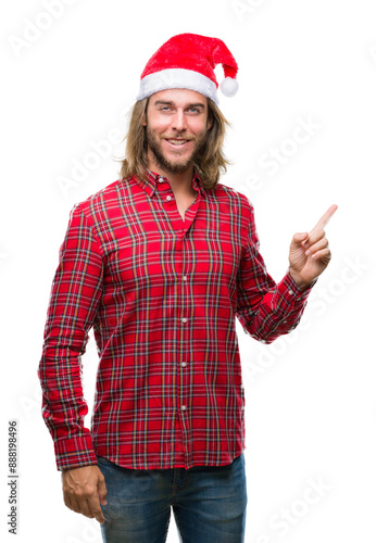 Young handsome man with long hair wearing santa claus hat over isolated background with a big smile on face, pointing with hand and finger to the side looking at the camera.