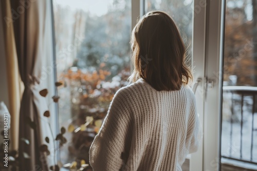 Woman in cozy sweater, looking out window at autumn scenery, morning sunlight, peaceful home atmosphere, reflection concept