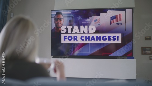 Adult woman sits on sofa at home with cup of coffee and watches political advertising or campaign about United States presidential elections on big TV screen. TV live broadcast. National Election Day.