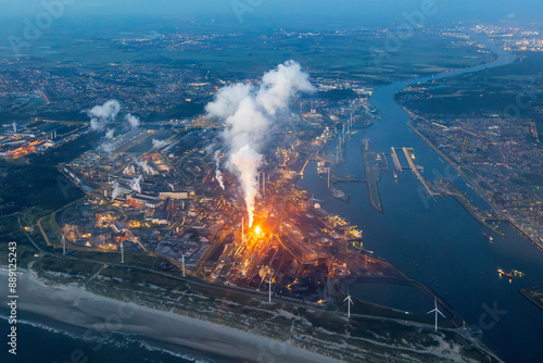 Aerial image of steel plant, factory, refinery with massive flame from chimney. Generating, producing steel and metal from coal and iron ore in IJmuiden Netherlands during evening alongside North Sea 