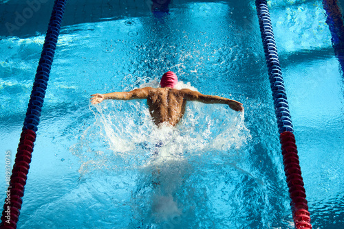 Swimmer, seen from above, powers through water with butterfly stroke, his muscular back glistening under sun as they swim between red lane dividers. Concept of aquatic sport, competition, energy.