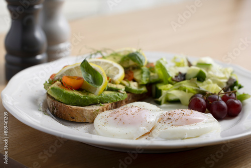 A delicious breakfast plate featuring poached eggs, avocado toast with lemon and tomato, a fresh salad, and a small bunch of grapes. Perfect for healthy eating and gourmet food themes.