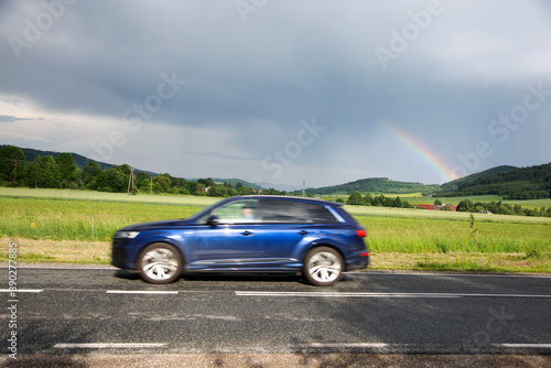 Samochód osobowy na drodze, tęcza nad górami, zielone pola, dachy domów.