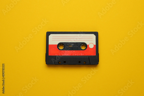 A cassette tape is laying on a yellow background. The tape is old and worn, with a red and white stripe. The tape is placed on a yellow surface, which gives the image a nostalgic and vintage feel