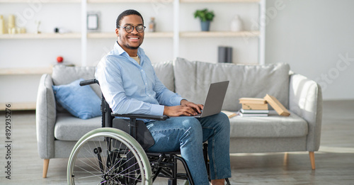 Happy disabled black man in wheelchair using laptop, working online from home, copy space. Millennial African American handicapped guy having business or studying on web