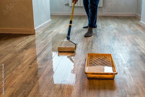 Worker varnishing floor with Eco white acrylic varnish for wood parquet floor, lacquering roller. Industrial theme. Home renovation